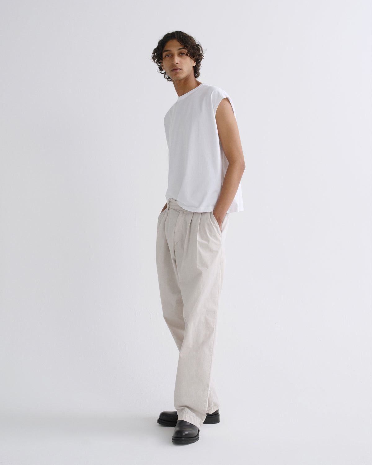 A model wearing a white sleeveless t-shirt and light beige linen trousers, standing against a plain light gray studio background.