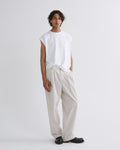 A model stands against a light gray studio background wearing a white sleeveless t-shirt, cream-colored wide-leg trousers, and black leather shoes.