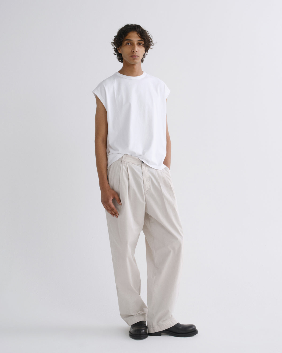 A model stands against a light gray studio background wearing a white sleeveless t-shirt, cream-colored wide-leg trousers, and black leather shoes.