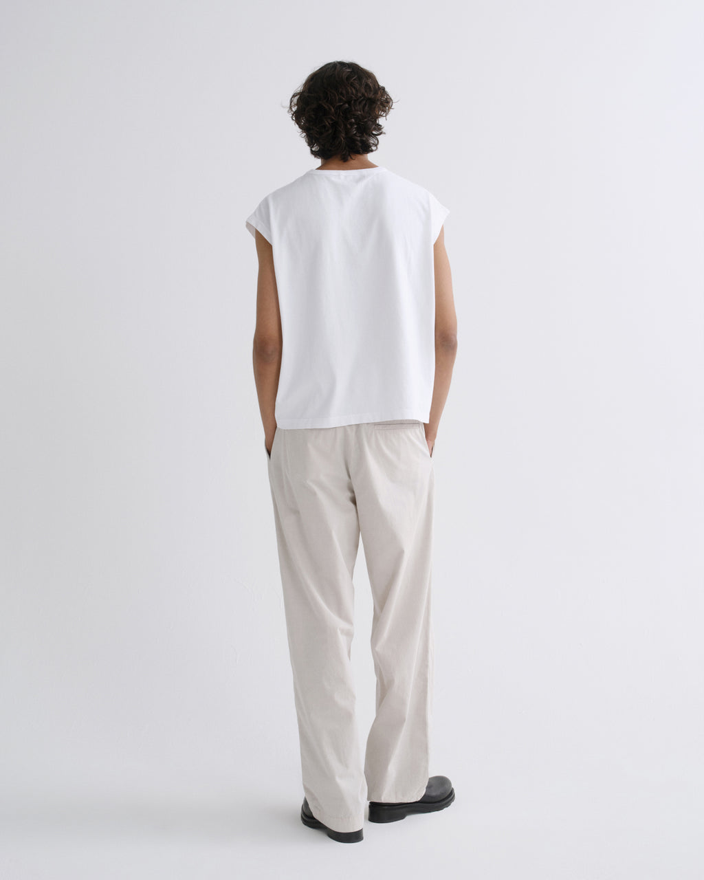 A model shown from the back wearing a white sleeveless t-shirt and light beige trousers against a plain light gray studio background.