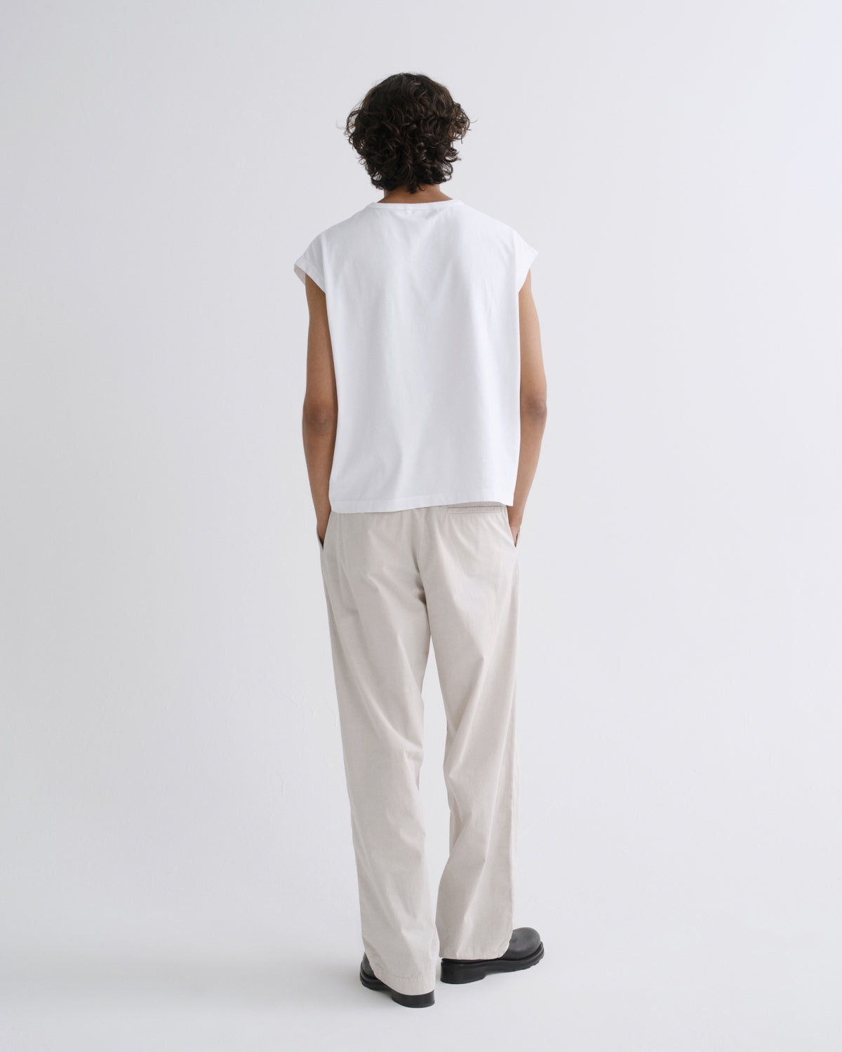 A model shown from the back wearing a white sleeveless t-shirt and light beige trousers against a plain light gray studio background.