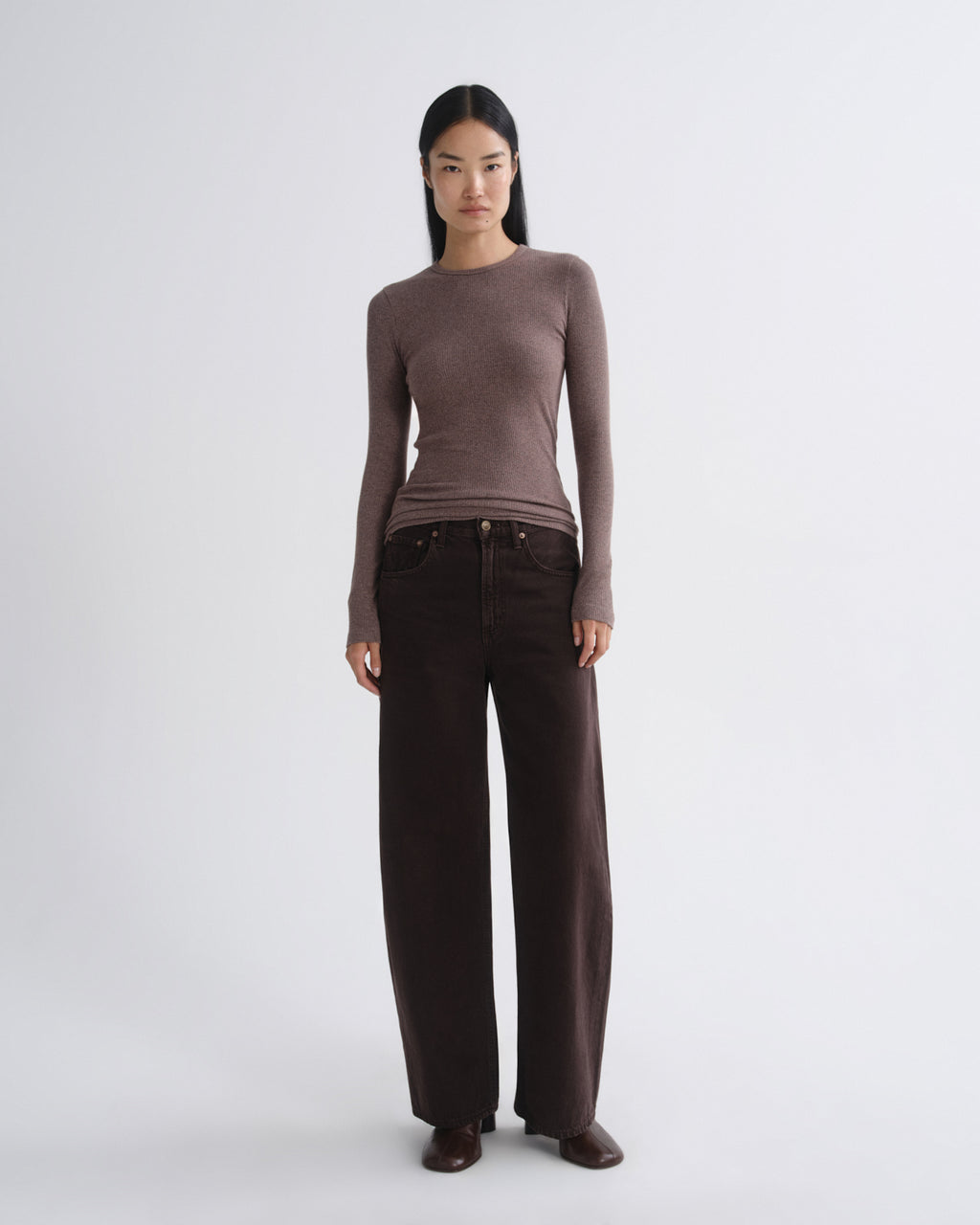 A model stands against a light gray studio background wearing a fitted long-sleeve crewneck top in taupe-brown and dark brown wide-leg trousers.