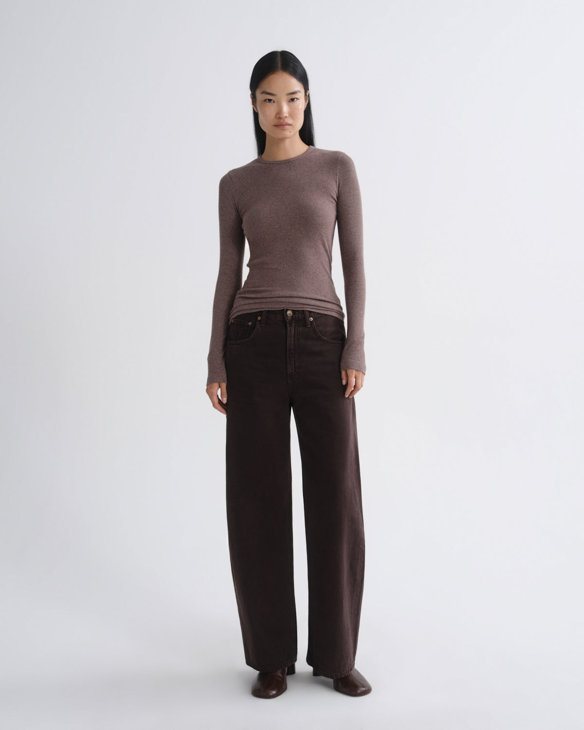 A model stands against a light gray studio background wearing a fitted long-sleeve crewneck top in taupe-brown and dark brown wide-leg trousers.