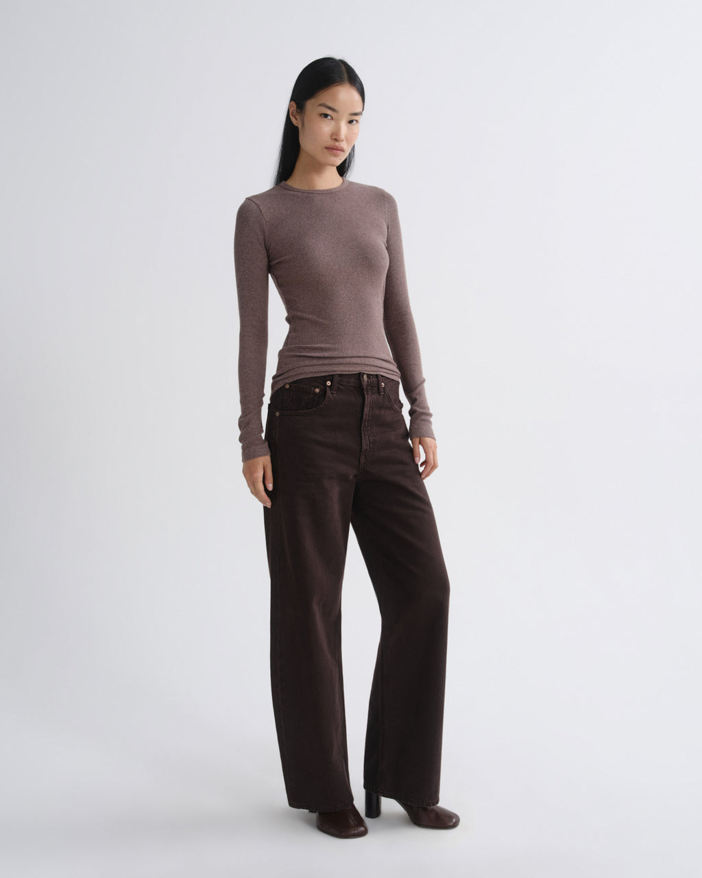 A woman modeling a fitted long-sleeve knit top in taupe-brown paired with dark brown wide-leg pants, standing against a light gray studio background.