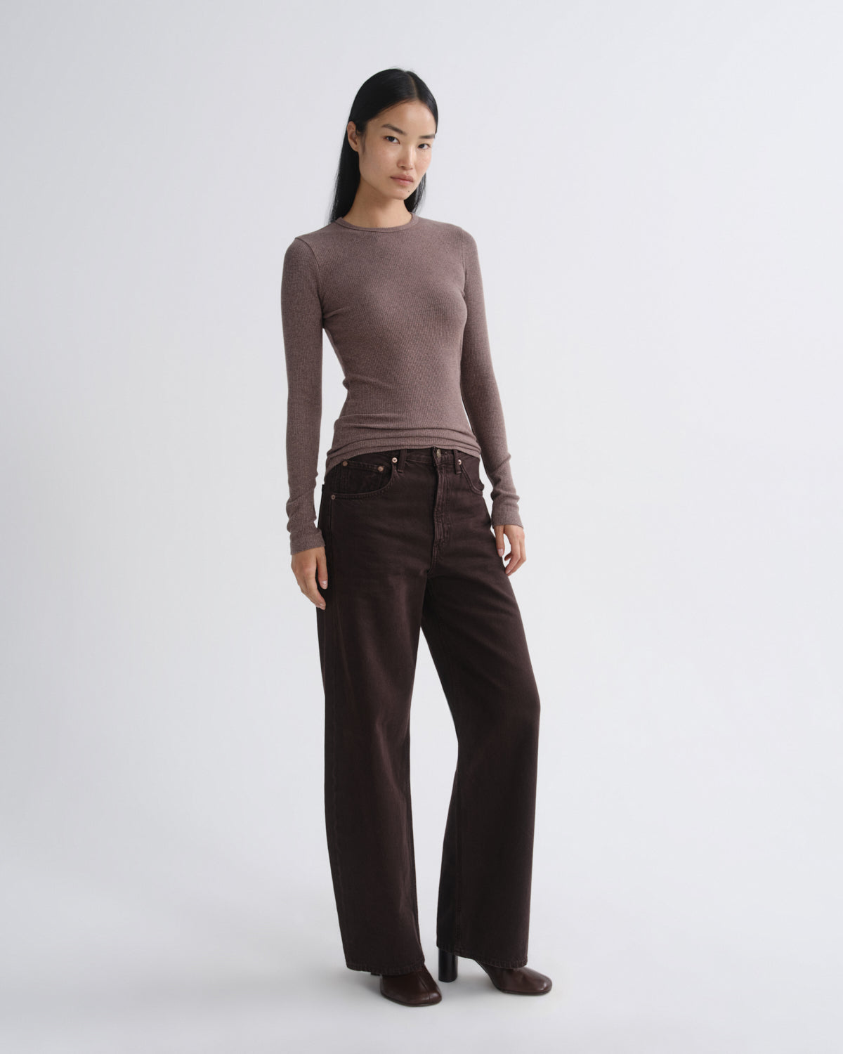 A woman modeling a fitted long-sleeve knit top in taupe-brown paired with dark brown wide-leg pants, standing against a light gray studio background.