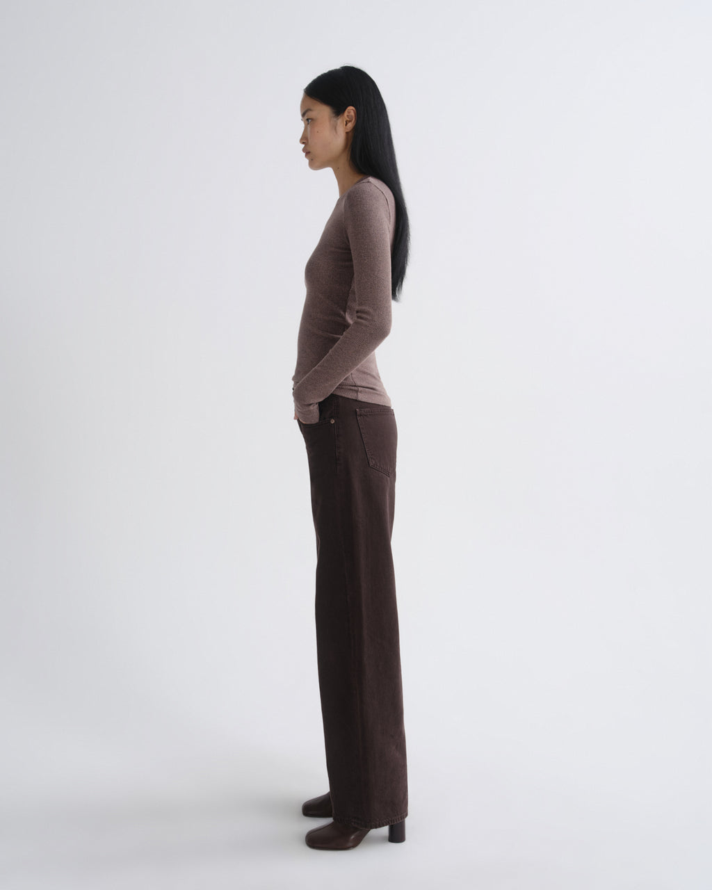 A woman stands in profile wearing a fitted long-sleeve top in a muted taupe-brown and dark brown wide-leg trousers, photographed against a clean light gray studio background.