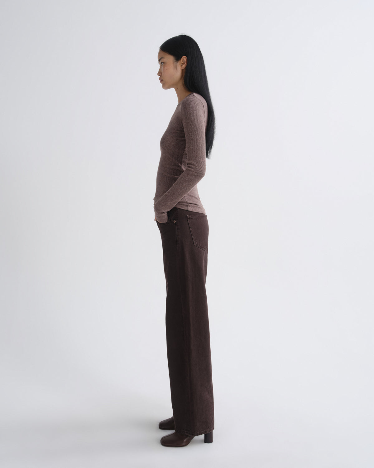 A woman stands in profile wearing a fitted long-sleeve top in a muted taupe-brown and dark brown wide-leg trousers, photographed against a clean light gray studio background.