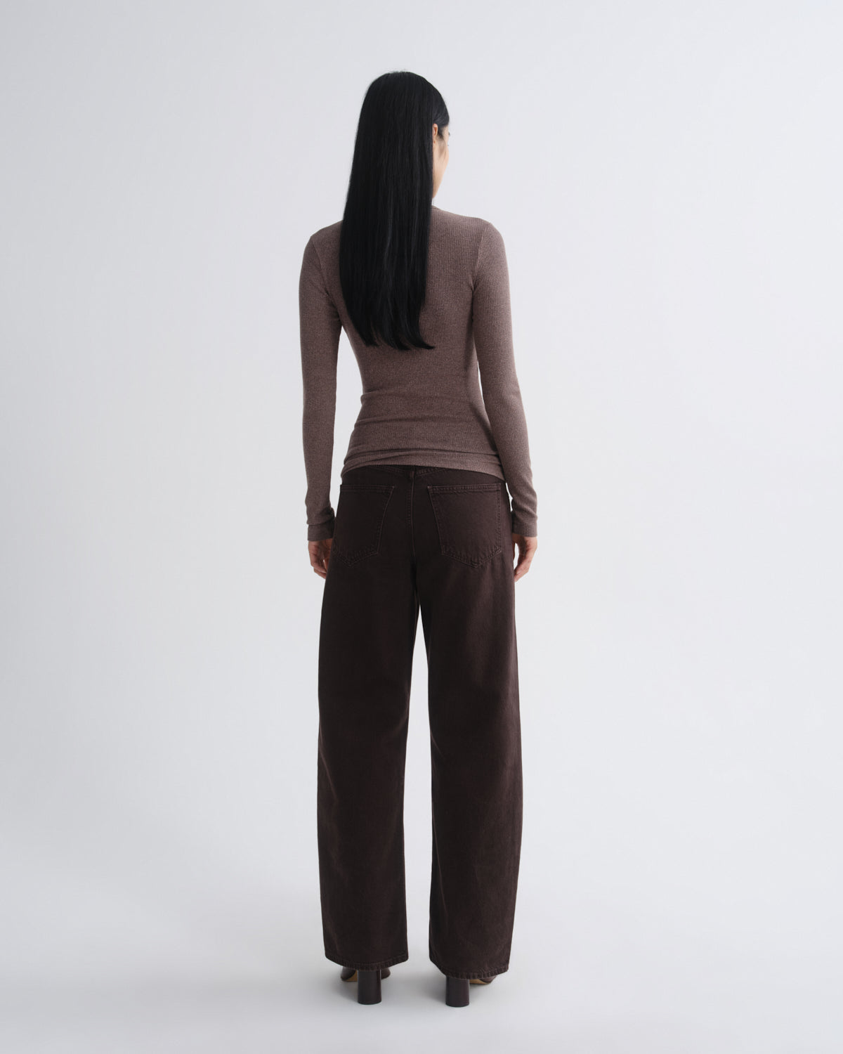 A model viewed from the back wearing a fitted taupe-brown long-sleeve top and dark brown wide-leg trousers, standing against a light gray studio background.