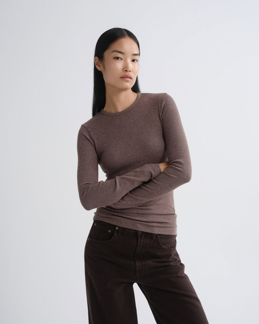 A woman modeling a fitted, long-sleeve crewneck knit top in a muted taupe-brown color, paired with dark brown pants, against a plain light gray studio background.