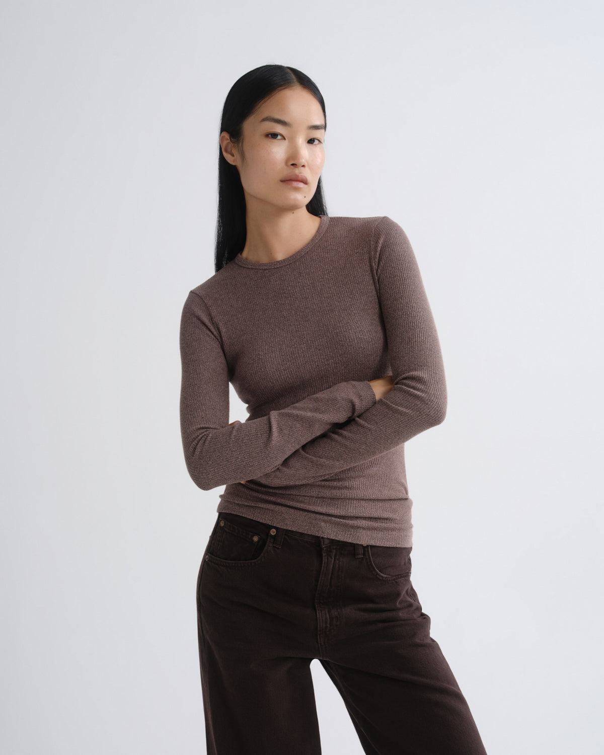 A woman modeling a fitted, long-sleeve crewneck knit top in a muted taupe-brown color, paired with dark brown pants, against a plain light gray studio background.