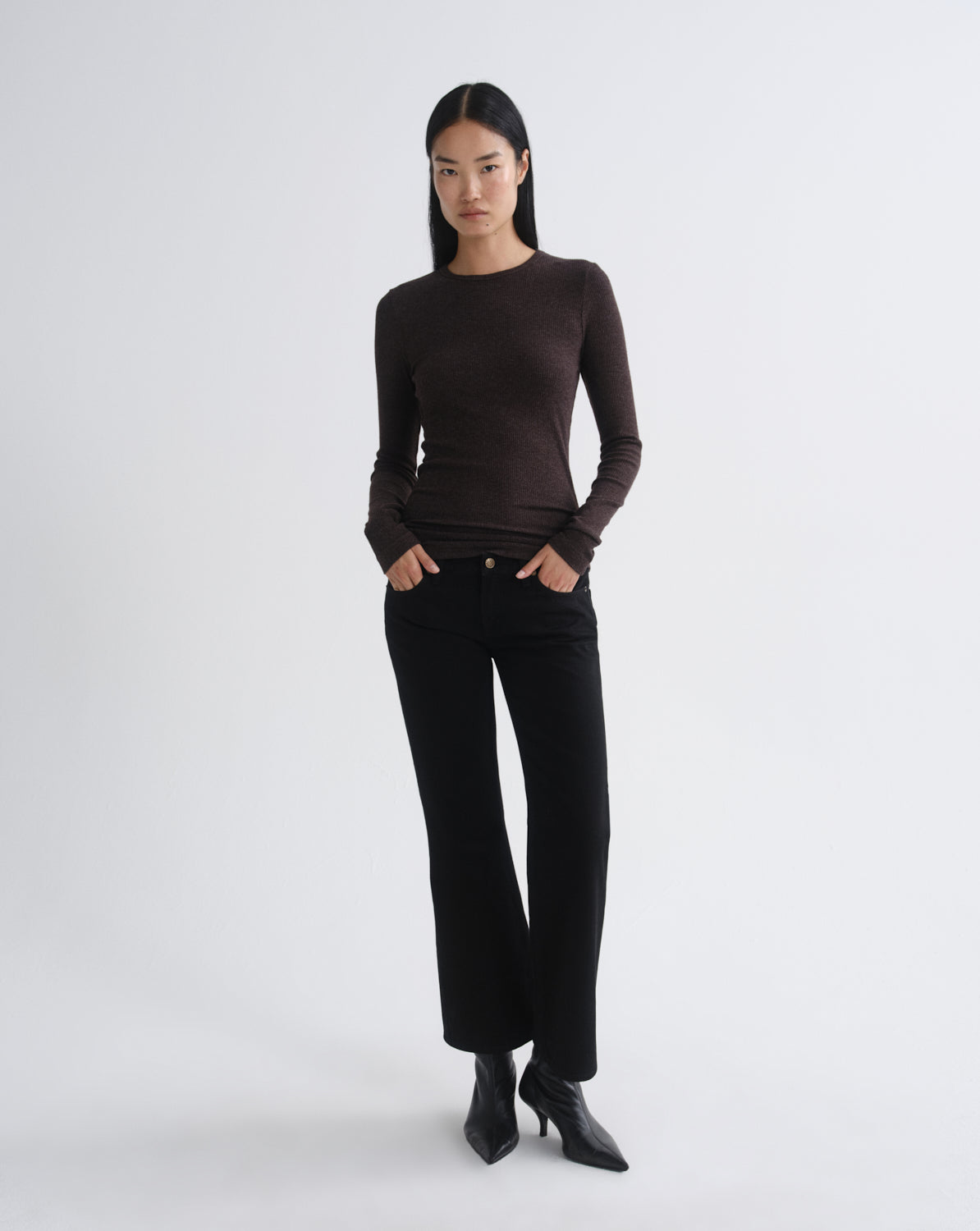 A model stands against a light gray studio background wearing a fitted dark brown long-sleeve top, black pants, and black pointed ankle boots.
