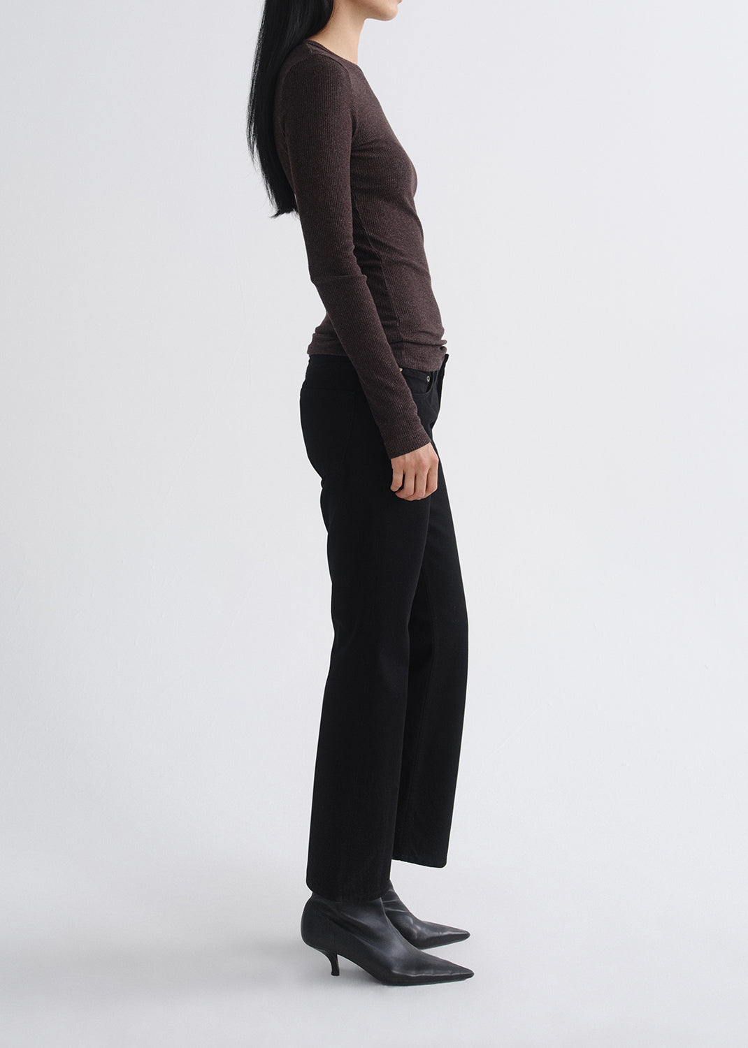 A side-profile studio shot of a model wearing a dark brown long-sleeve knit top, black wide-leg trousers, and black pointed-toe ankle boots against a grey  background.