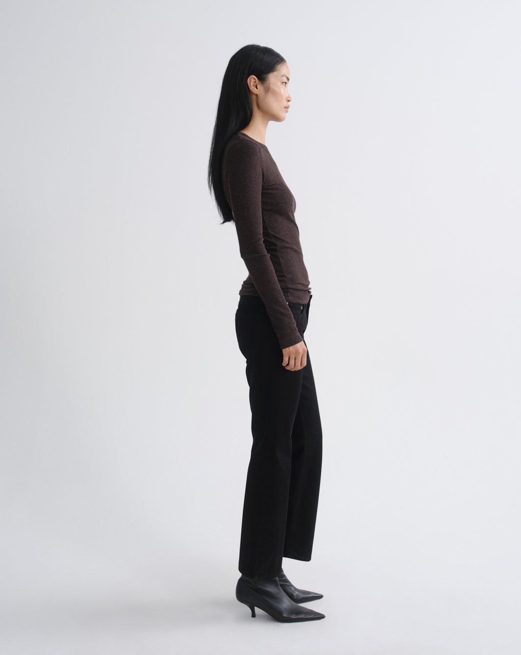 A woman in side profile wearing a dark brown long-sleeve top, black wide-leg trousers, and black ankle boots, standing against a light gray studio background.