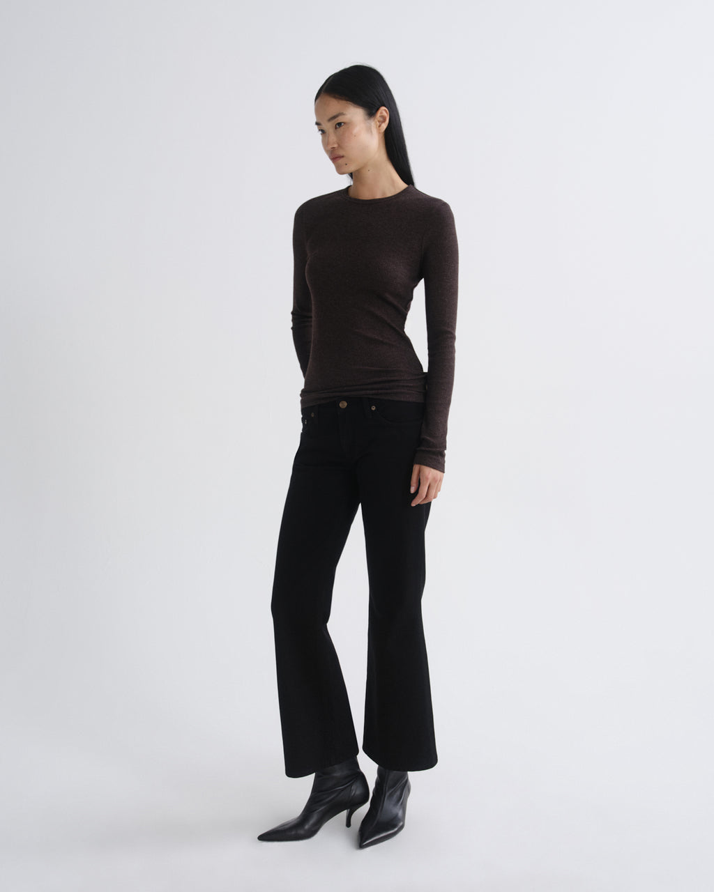 A model wearing a dark brown long-sleeve crewneck top, black wide-leg cropped pants, and black pointed-toe ankle boots, standing against a light gray studio background.