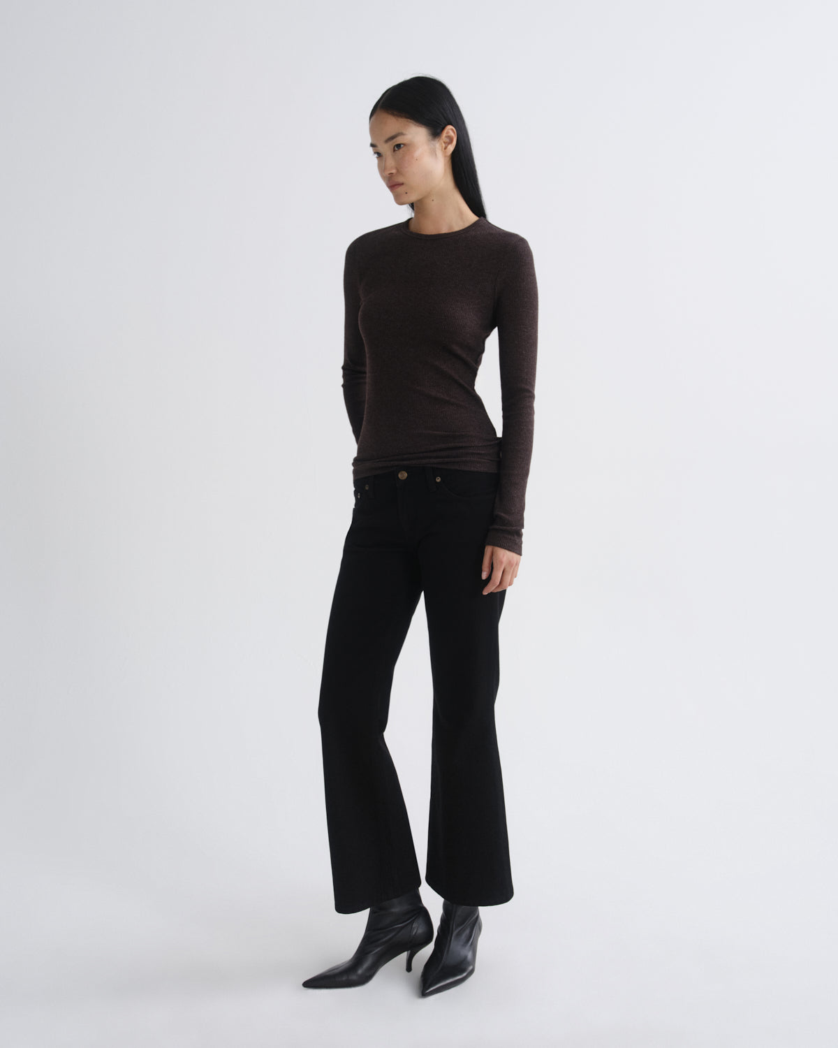 A model wearing a dark brown long-sleeve crewneck top, black wide-leg cropped pants, and black pointed-toe ankle boots, standing against a light gray studio background.