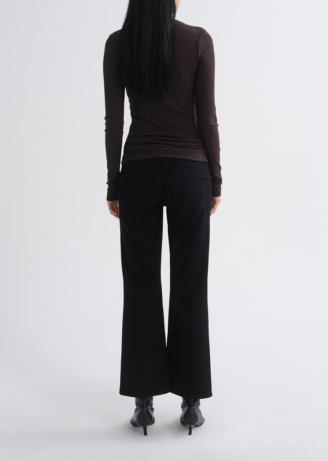 A model shown from the back wearing a dark brown long-sleeve top, black wide-leg pants, and black pointed-toe heels against a grey background.