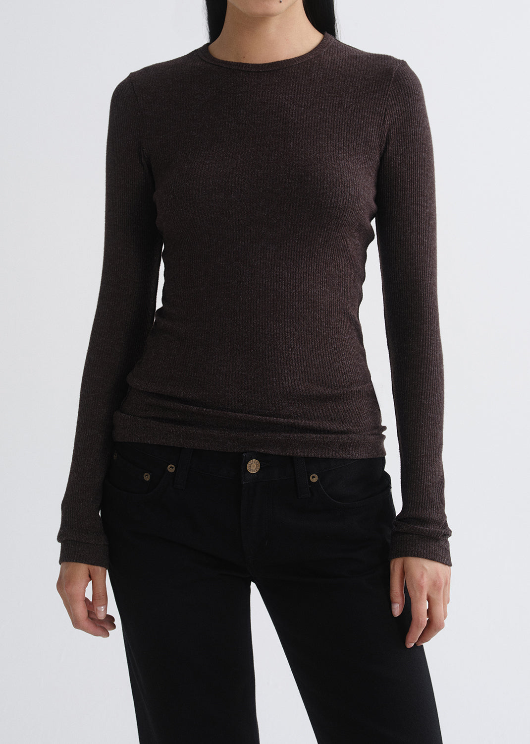 A woman wearing a dark brown ribbed long-sleeve crewneck sweater and black jeans, shown from the neck down against a grey background.