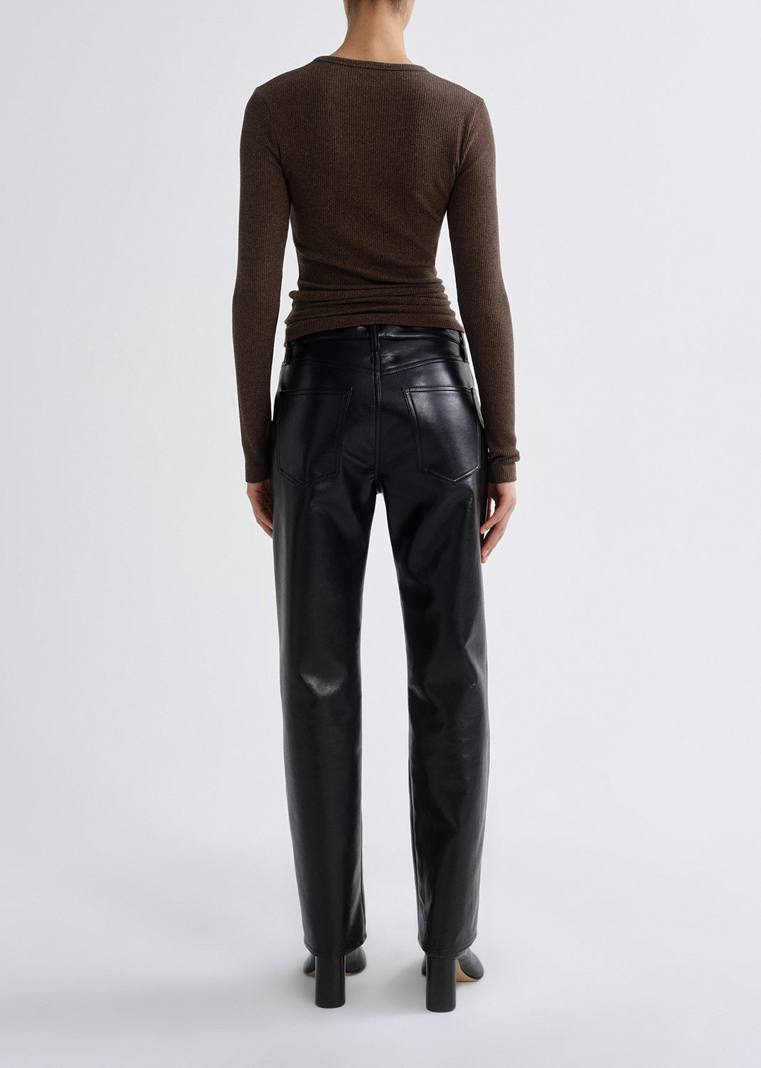 Person wearing a brown long sleeve ribbed top with black leather pants viewed from the back on a grey background.