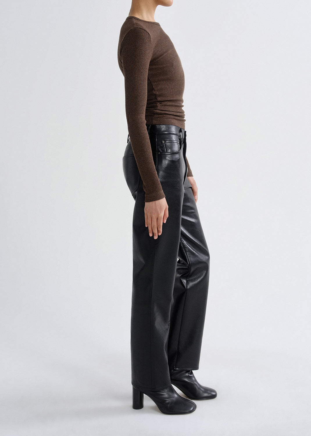 Person wearing a brown long sleeve ribbed top with black leather pants viewed from the side on a grey background.