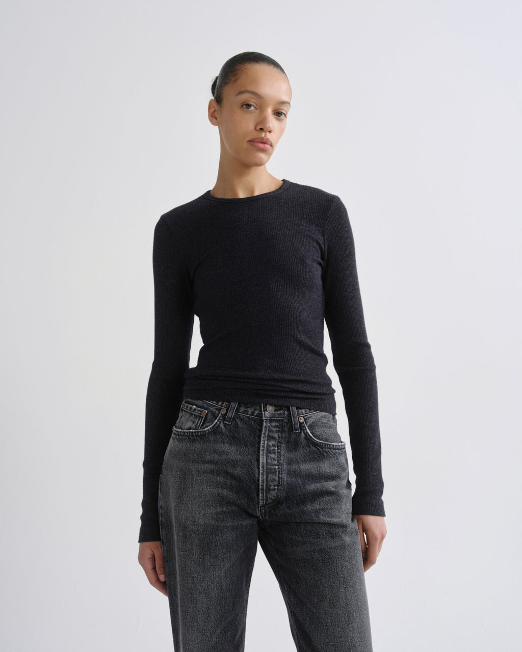 A model wearing a dark charcoal long-sleeve crewneck sweater and dark gray jeans, standing against a light gray studio background.