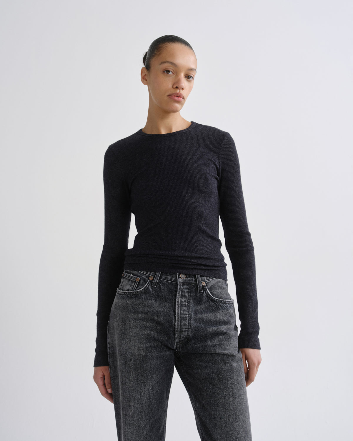 A model wearing a dark charcoal long-sleeve crewneck sweater and dark gray jeans, standing against a light gray studio background.