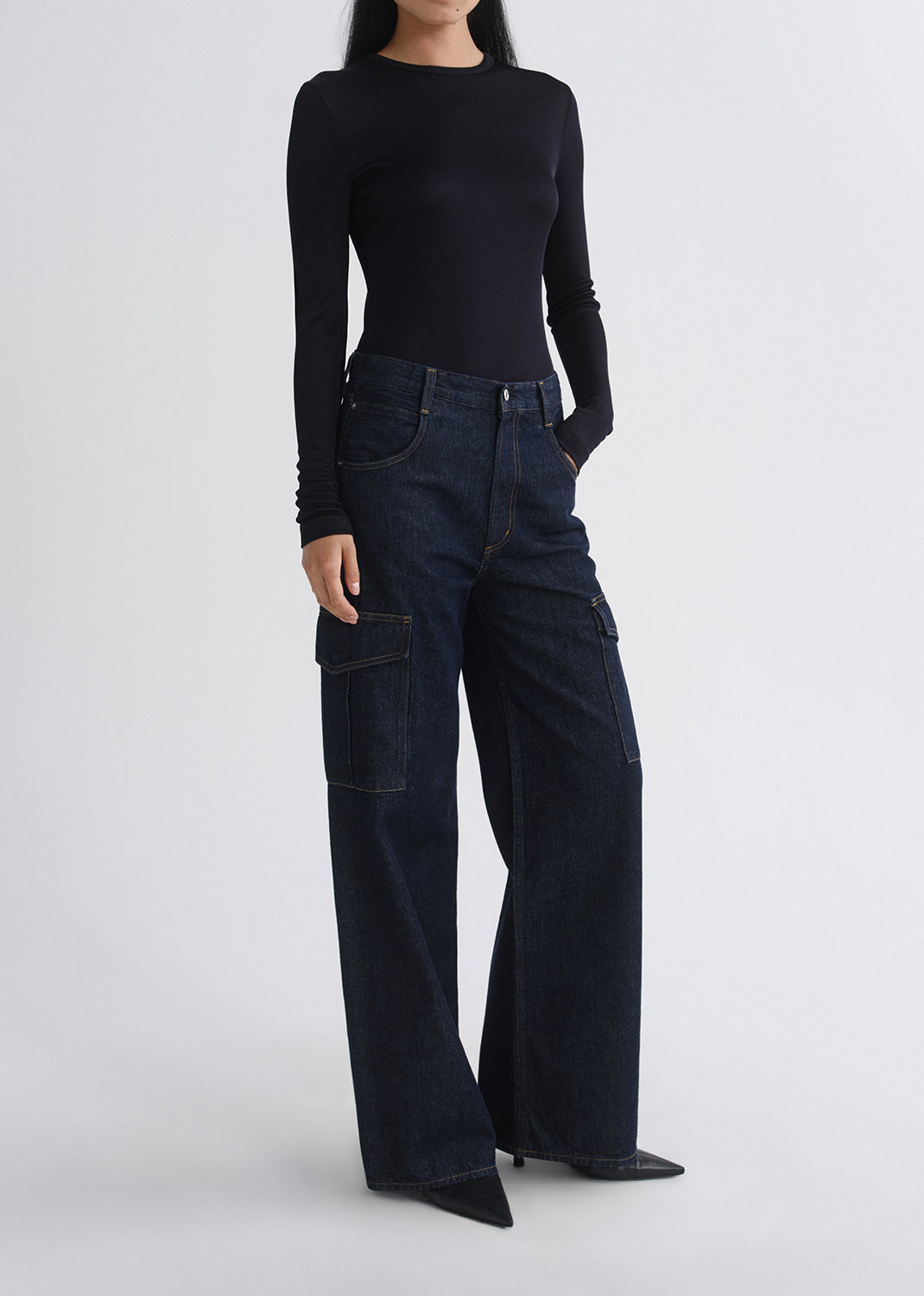 A model wearing a black long-sleeve top and dark wide-leg cargo jeans with side flap pockets, standing against a grey background.