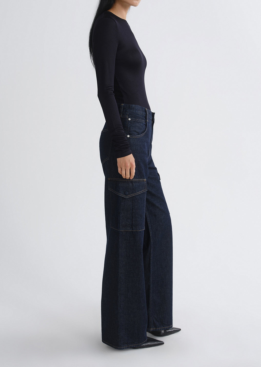 A side-profile studio shot of a model wearing dark indigo wide-leg denim jeans with front cargo pockets and a black long-sleeve top, set against a grey background.