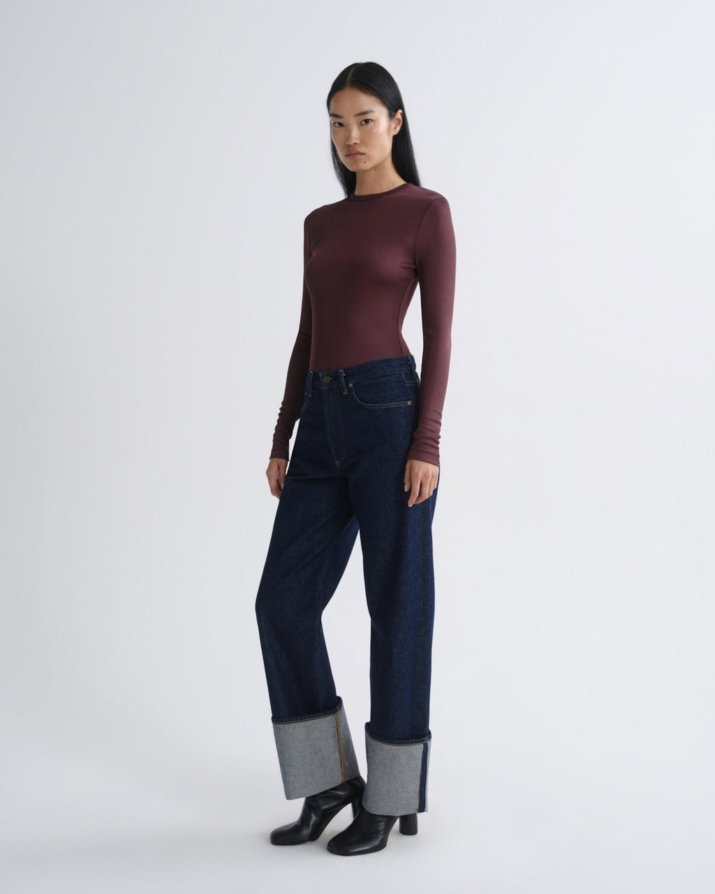 Woman wearing a burgundy long-sleeve top and dark blue jeans with black boots on a white background