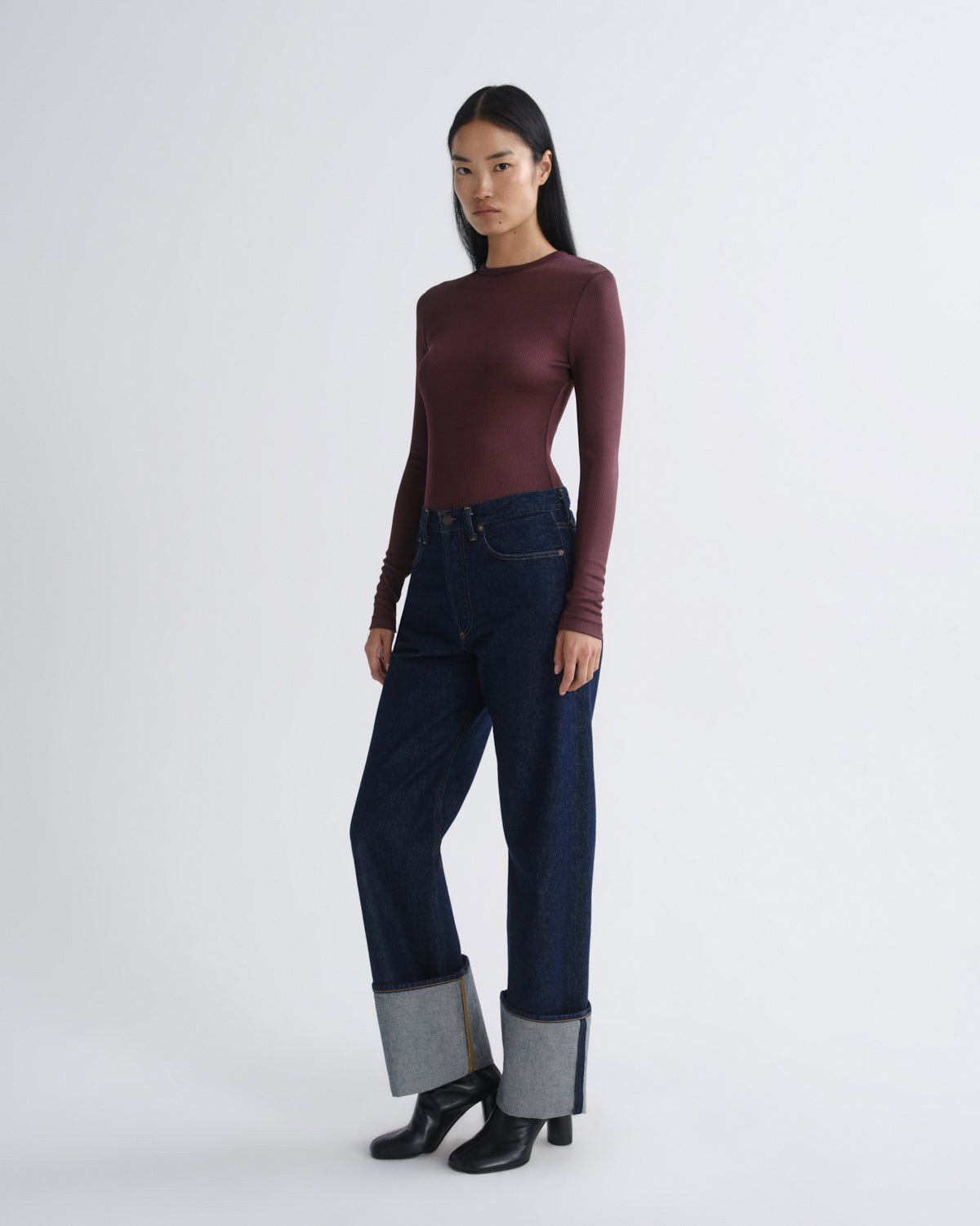 Woman wearing a burgundy long-sleeve top and dark blue jeans with black boots on a white background