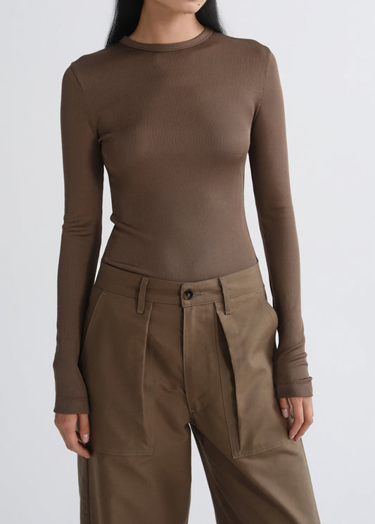 A model wearing a fitted, long-sleeve crewneck top in a warm taupe-brown color, paired with high-waisted brown pleated trousers, against a grey background.