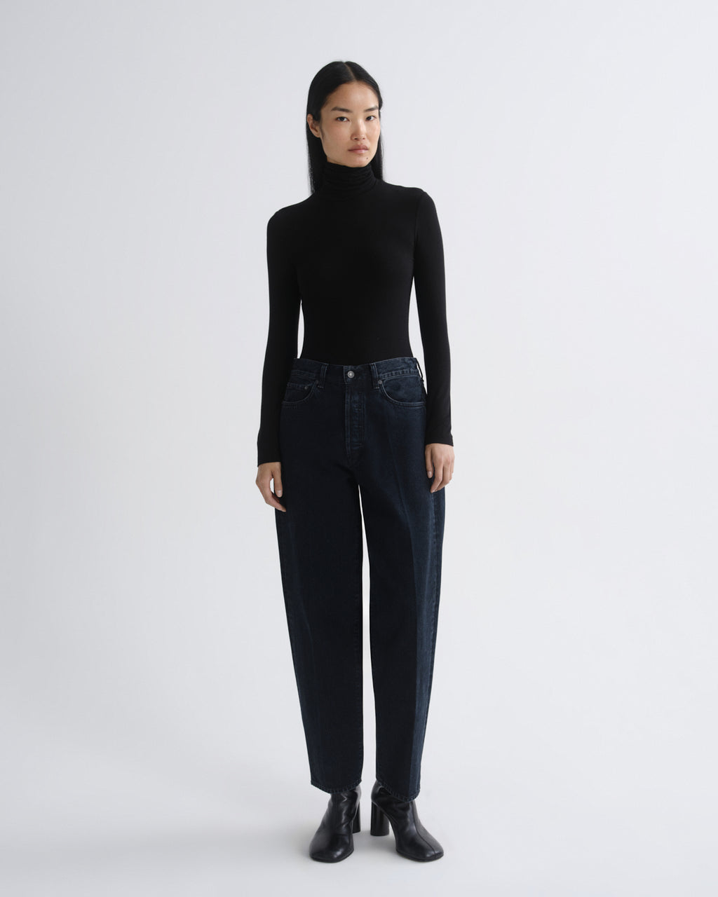 A model stands against a light gray studio background wearing a black long-sleeve turtleneck sweater, dark blue high-waisted straight-leg jeans, and black ankle boots.