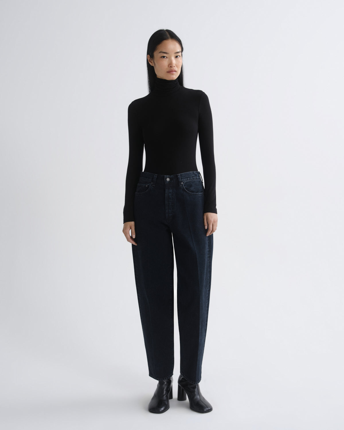 A model stands against a light gray studio background wearing a black long-sleeve turtleneck sweater, dark blue high-waisted straight-leg jeans, and black ankle boots.