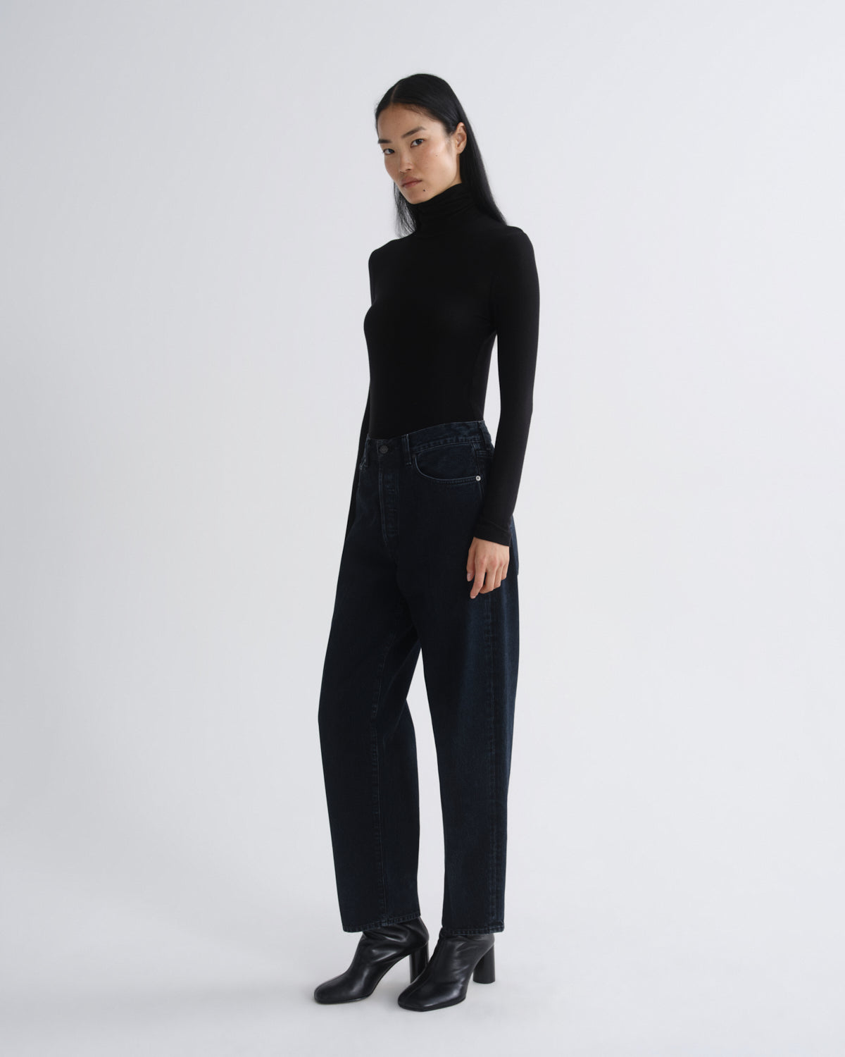 A woman modeling a black long-sleeve mock neck top and dark wide-leg jeans with black heeled ankle boots, standing against a light gray studio background.