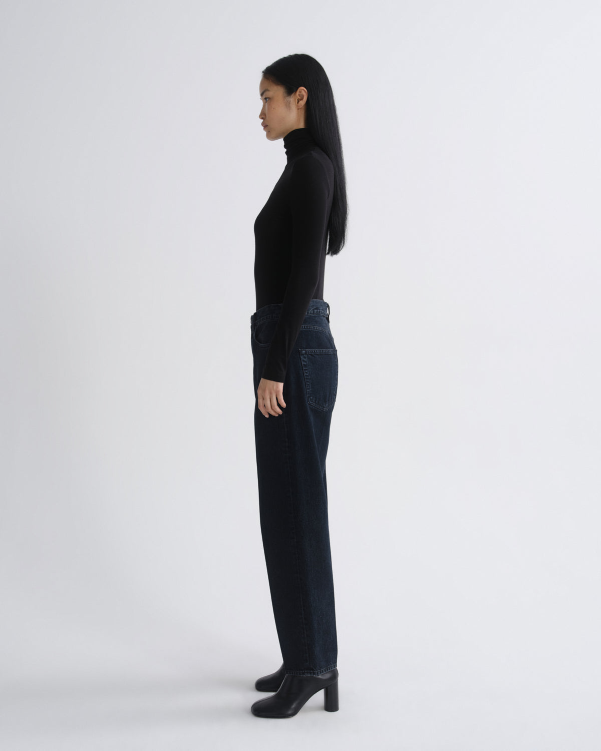 A woman shown from the back wearing a black long-sleeve top, dark blue straight-leg jeans, and black block-heel boots against a light gray studio background.