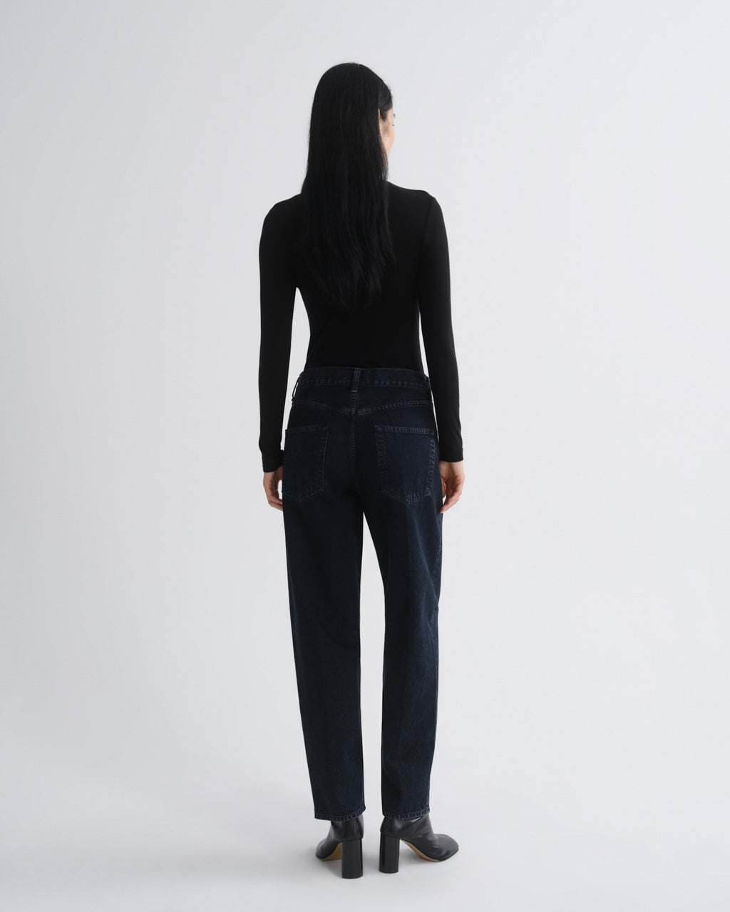 A woman shown from the back wearing a black long-sleeve top, dark blue straight-leg jeans, and black block-heel boots against a light gray studio background.