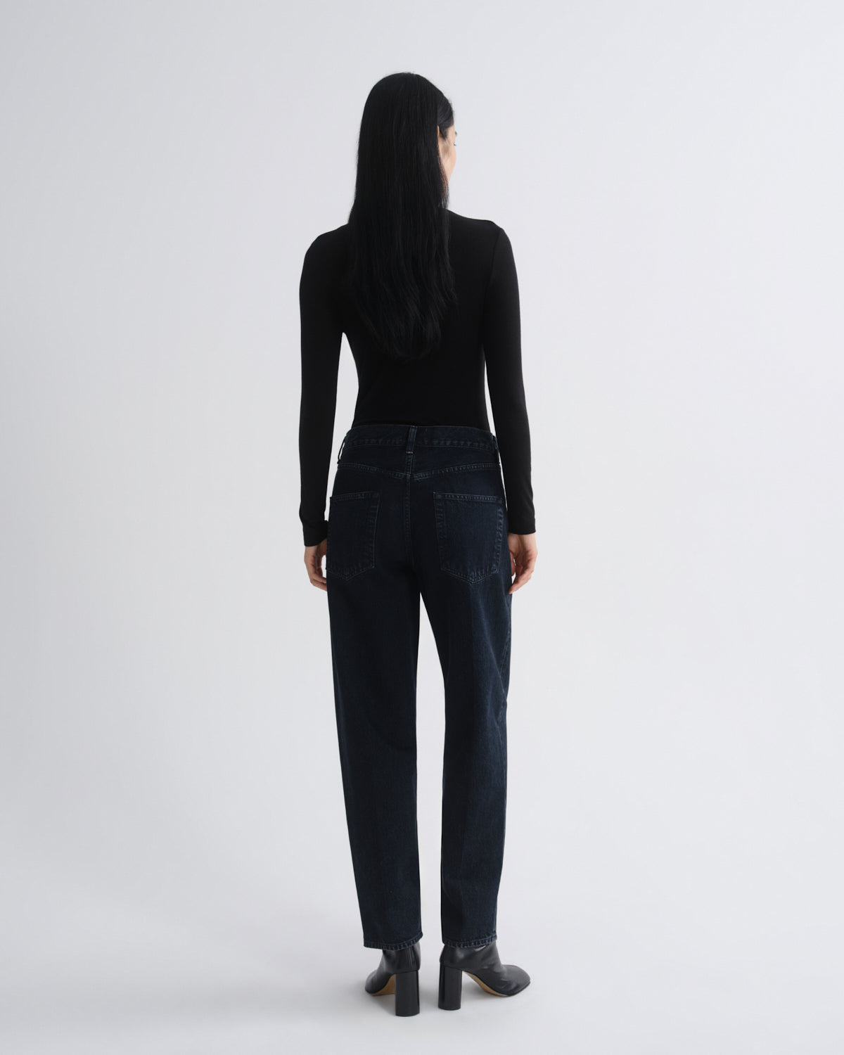 A woman shown from the back wearing a black long-sleeve top, dark blue straight-leg jeans, and black block-heel boots against a light gray studio background.