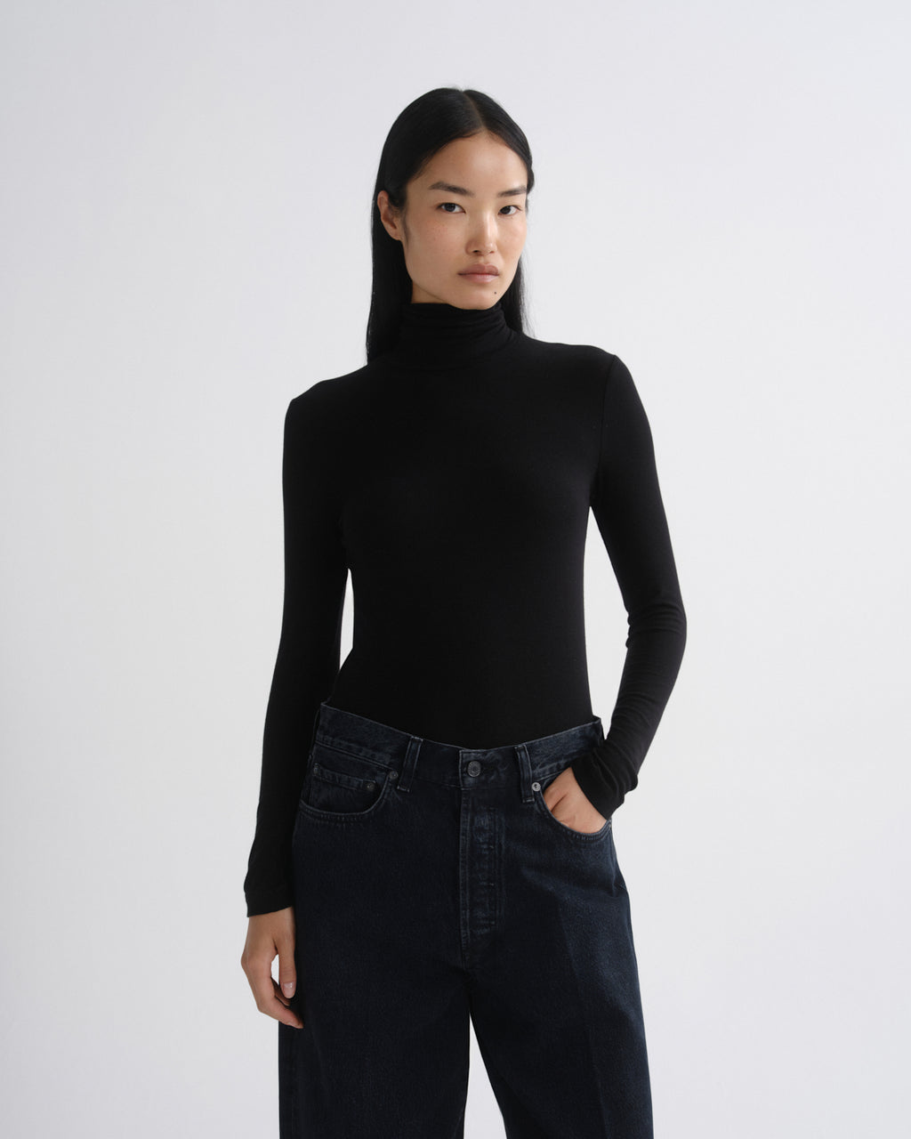 A model wearing a black long-sleeve mock neck turtleneck top and dark blue jeans, standing against a light gray studio background.
