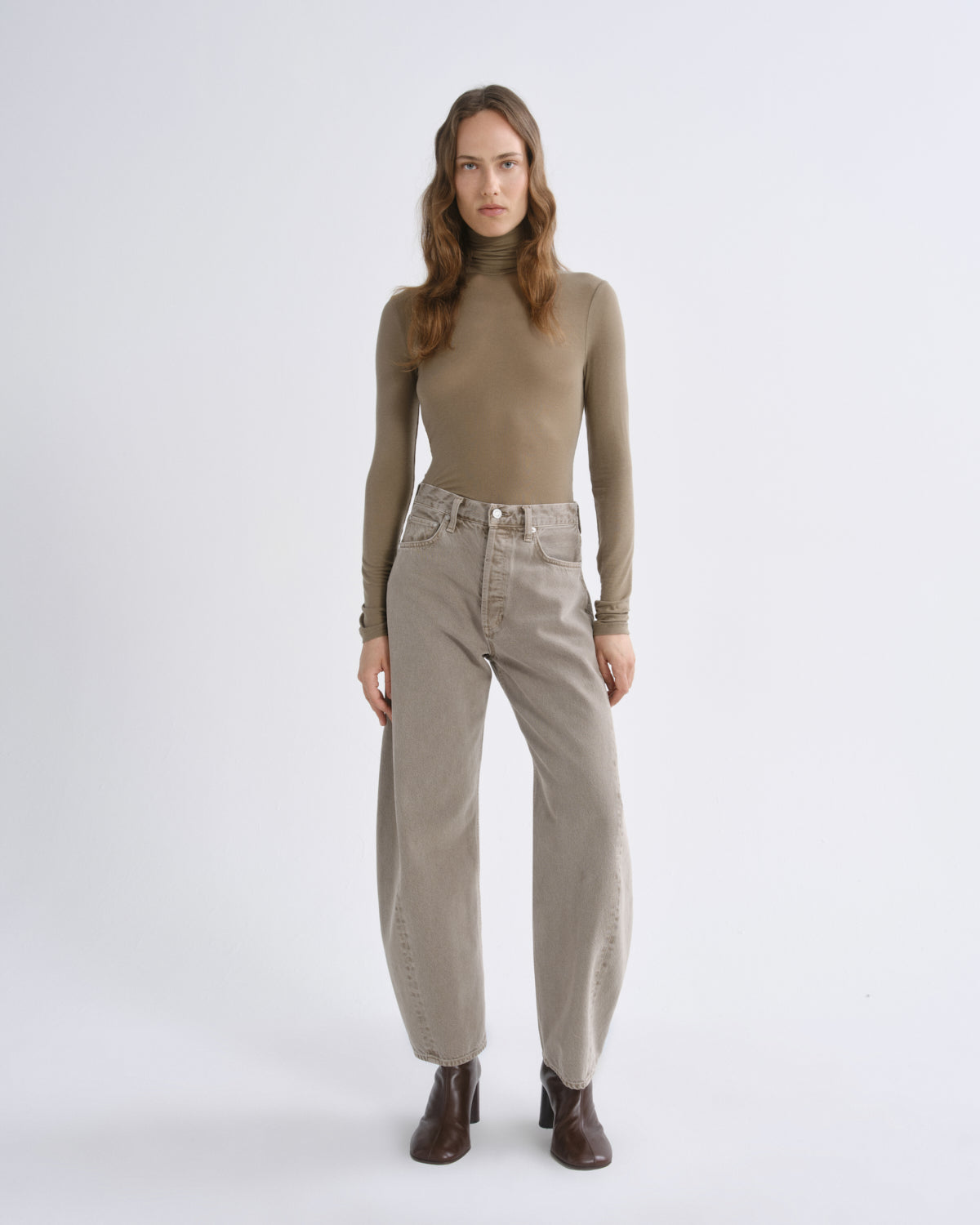 A woman stands against a light gray studio background wearing a fitted taupe mock-neck long-sleeve top, light beige wide-leg jeans, and dark brown ankle boots.