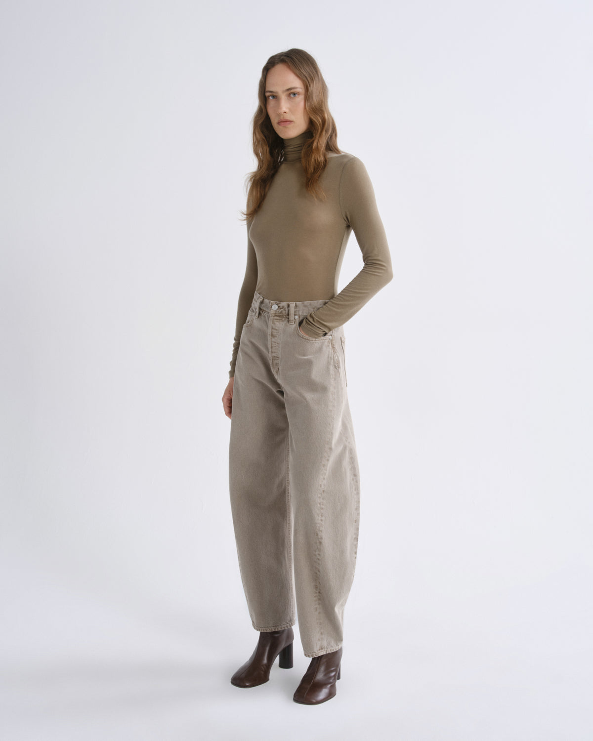A woman modeling a fitted taupe long-sleeve mock-neck top and wide-leg beige trousers, paired with dark brown ankle boots, against a plain light gray studio background.
