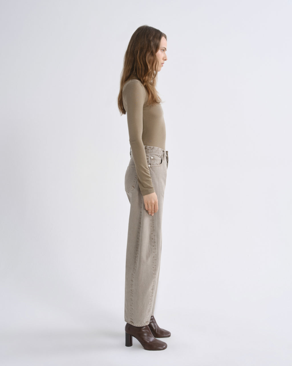 A woman stands in profile wearing a fitted long-sleeve top, wide-leg cropped pants, and dark brown heeled ankle boots against a plain light gray studio background.