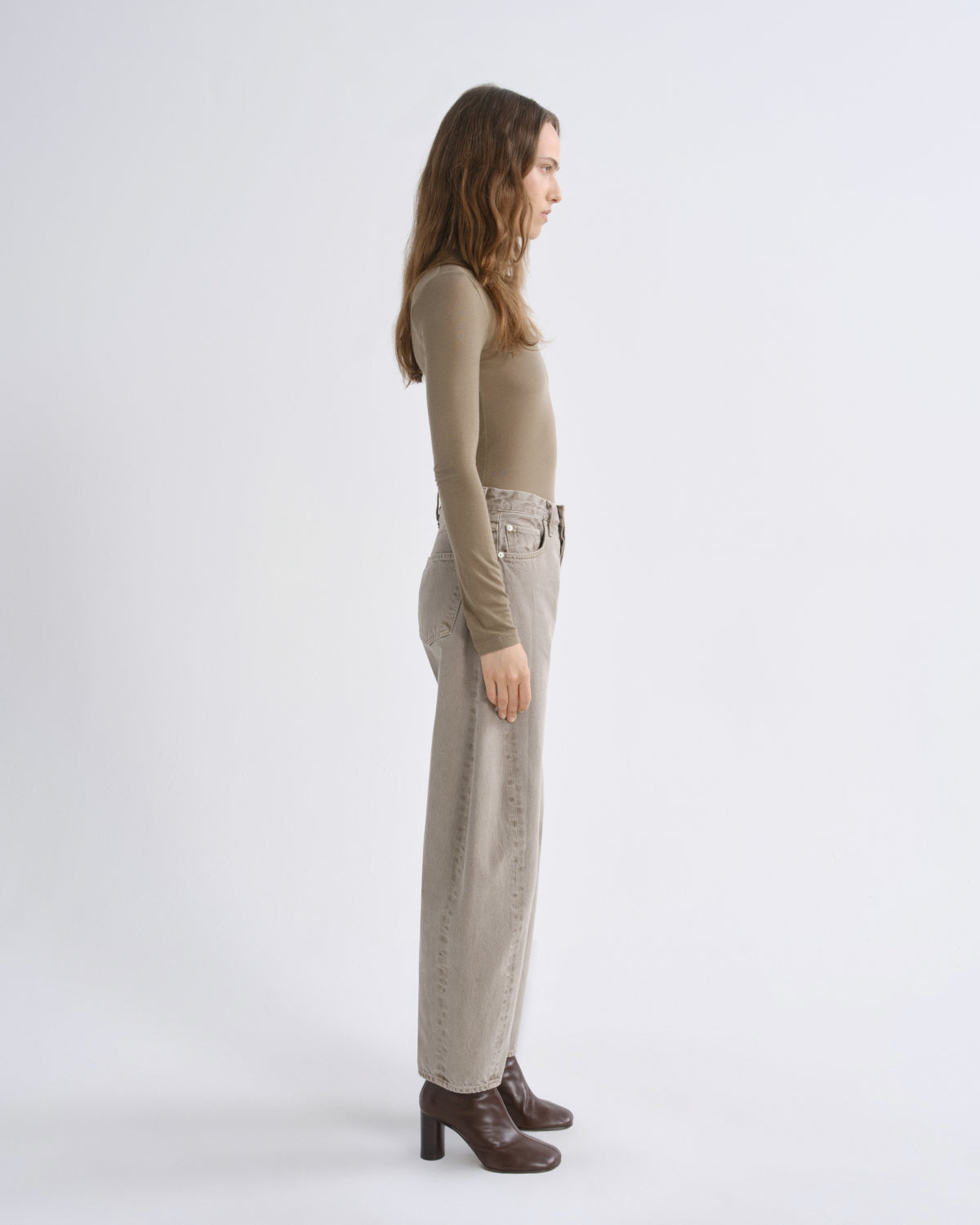A woman stands in profile wearing a fitted long-sleeve top, wide-leg cropped pants, and dark brown heeled ankle boots against a plain light gray studio background.
