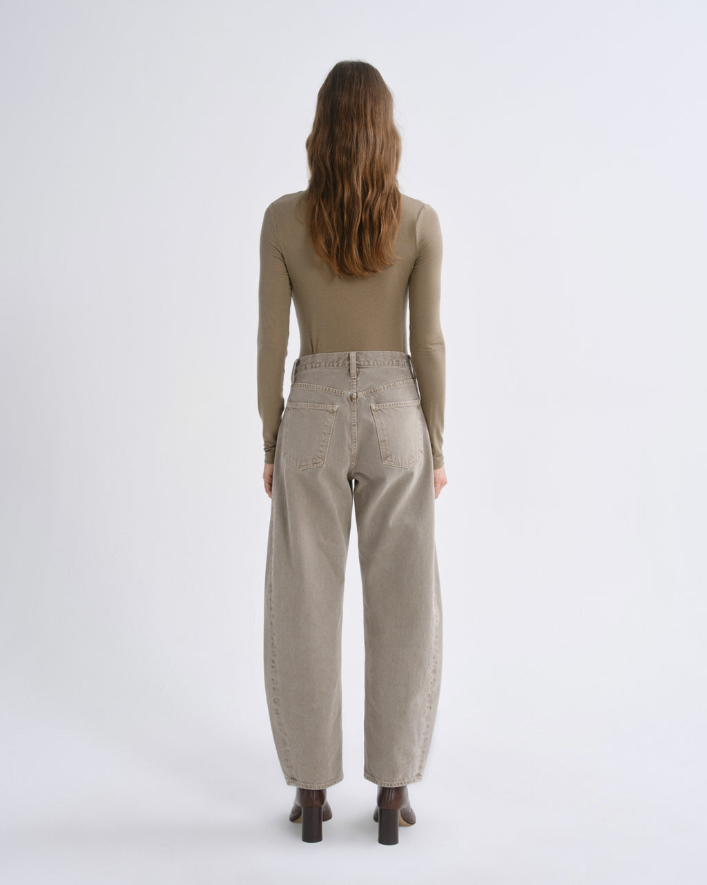A woman viewed from behind wearing a fitted taupe long-sleeve top and wide-leg beige trousers, standing against a plain light gray studio background.