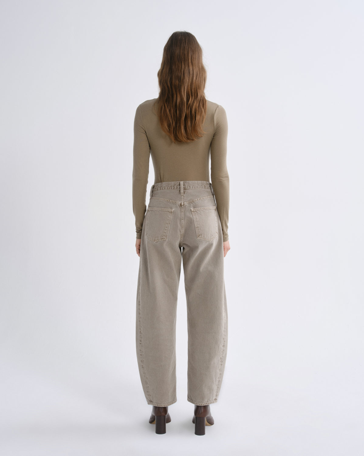 A woman viewed from behind wearing a fitted taupe long-sleeve top and wide-leg beige trousers, standing against a plain light gray studio background.
