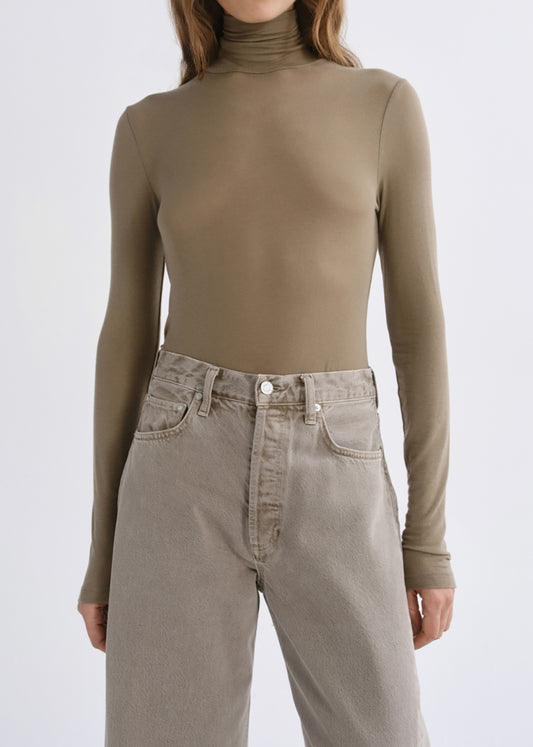 A model wearing a fitted, long-sleeve ribbed turtleneck top in a muted taupe-brown color, paired with light-wash beige jeans, against a grey background.