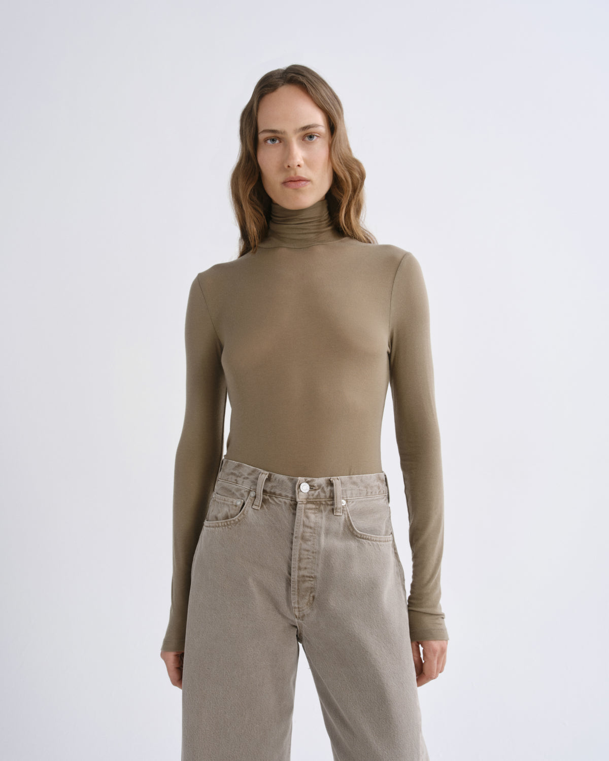 A woman modeling a fitted, long-sleeve ribbed turtleneck top in a muted taupe-brown color, paired with light beige jeans, against a plain light gray studio background.