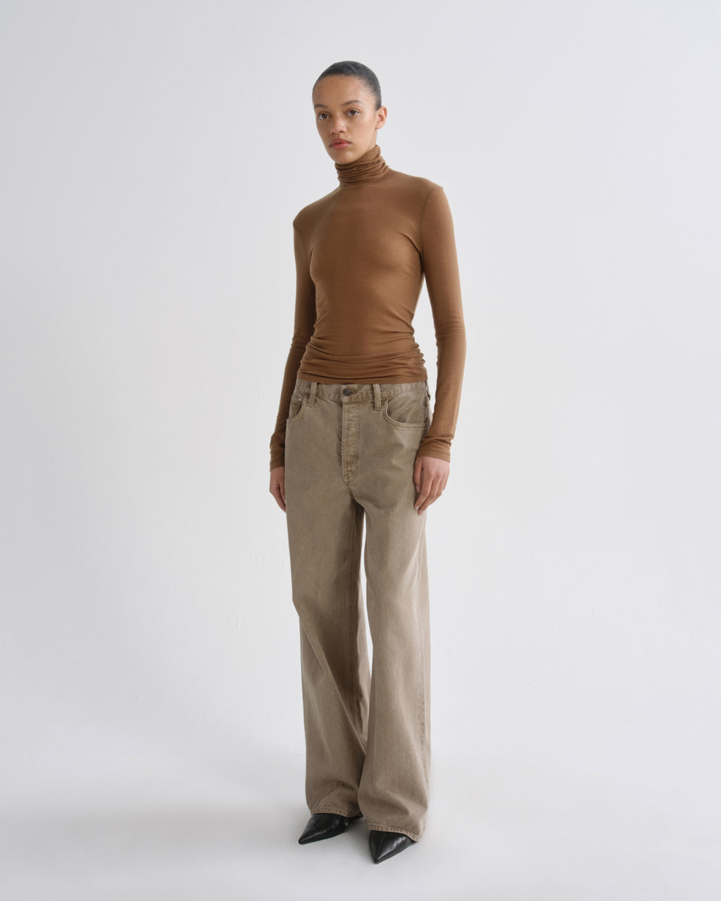 A model stands against a light gray studio background wearing a fitted brown turtleneck top, wide-leg beige pants, and black pointed shoes.