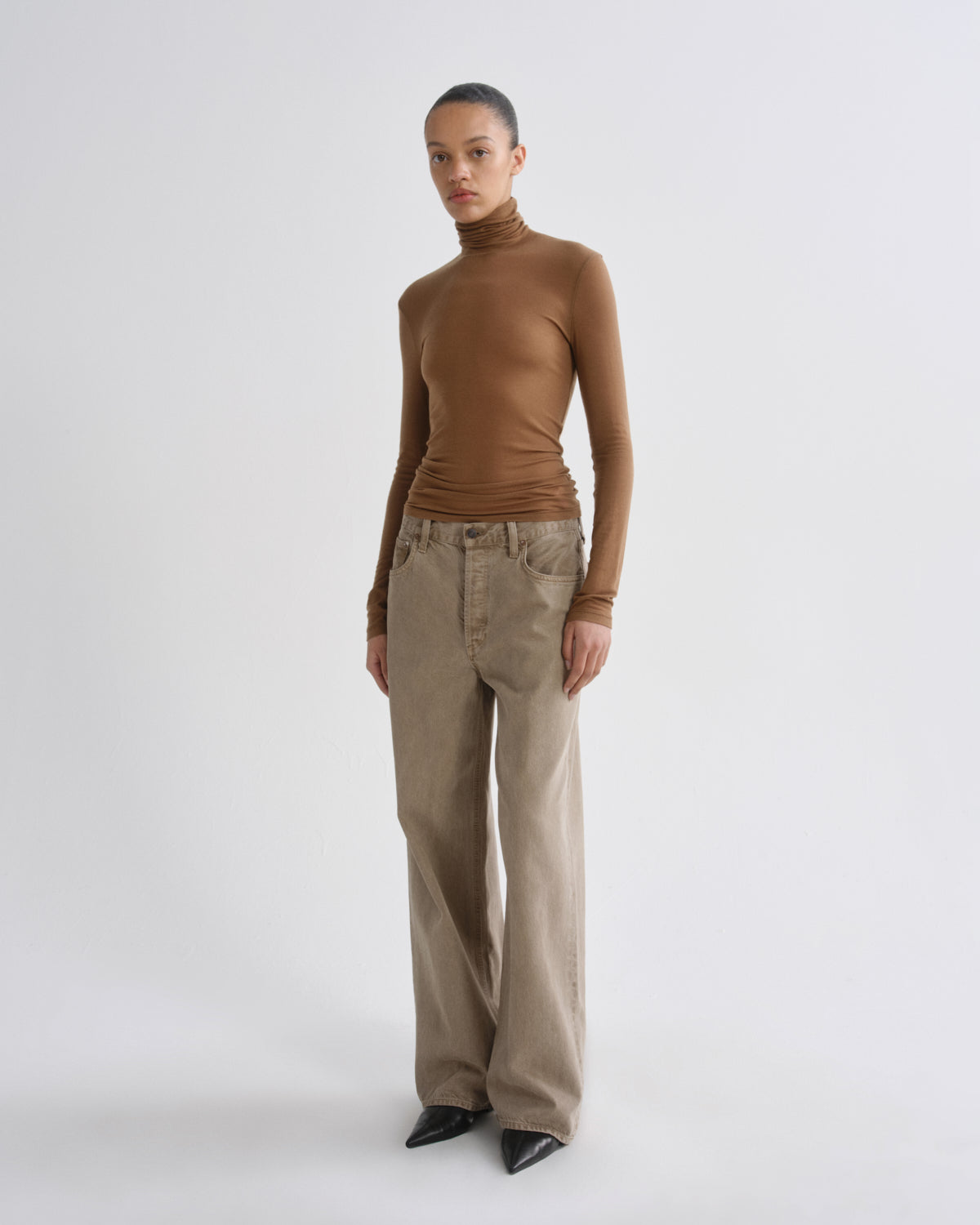 A model stands against a light gray studio background wearing a fitted brown turtleneck top, wide-leg beige pants, and black pointed shoes.