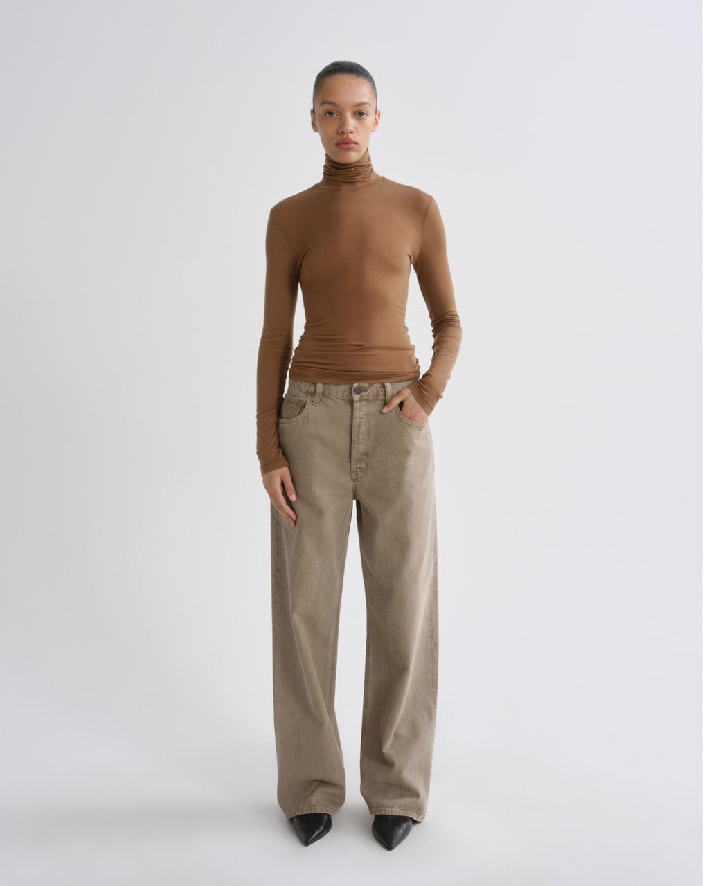 A model stands against a light gray studio background wearing a fitted brown ribbed turtleneck top and wide-leg beige trousers with black pointed shoes.