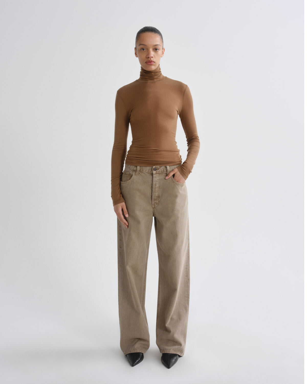 A model stands against a light gray studio background wearing a fitted brown ribbed turtleneck top and wide-leg beige trousers with black pointed shoes.