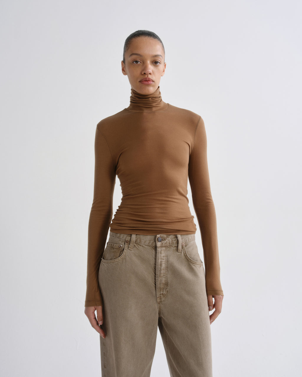 A model wearing a fitted, long-sleeve brown ruffled mock neck top and beige high-waisted jeans, standing against a light gray studio background.
